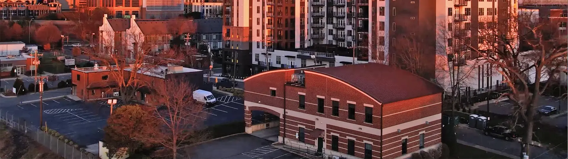 Aerial view of urban buildings, including a red brick structure with arched windows and a parking lot, set against a backdrop of modern multi-story apartments during sunset.