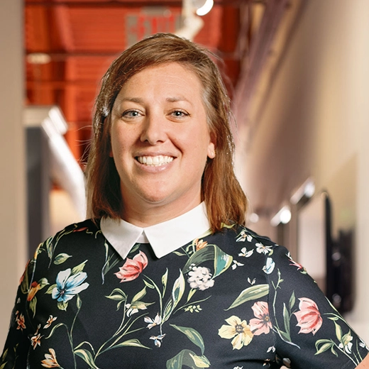 Smiling person with brown hair in a floral dress stands in a hallway with a red ceiling.