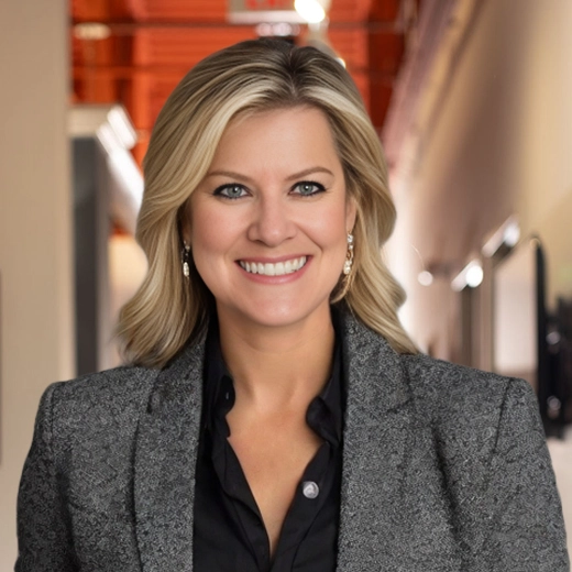 A person with blonde hair smiling, wearing a gray blazer and black shirt, standing in a well-lit hallway.