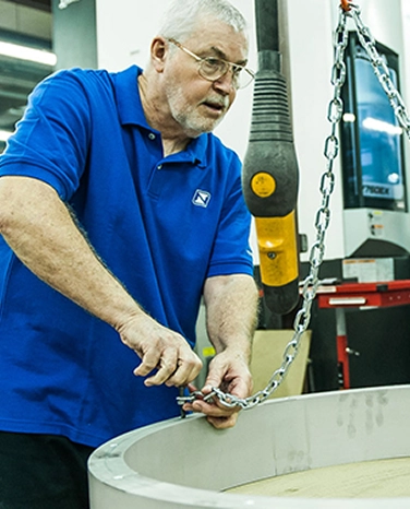 Older man in a blue shirt fixing a metal chain around a large circular object in a workshop.
