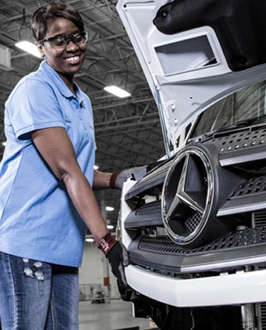 Person in a blue shirt working on a Mercedes-Benz vehicle hood in an industrial setting.