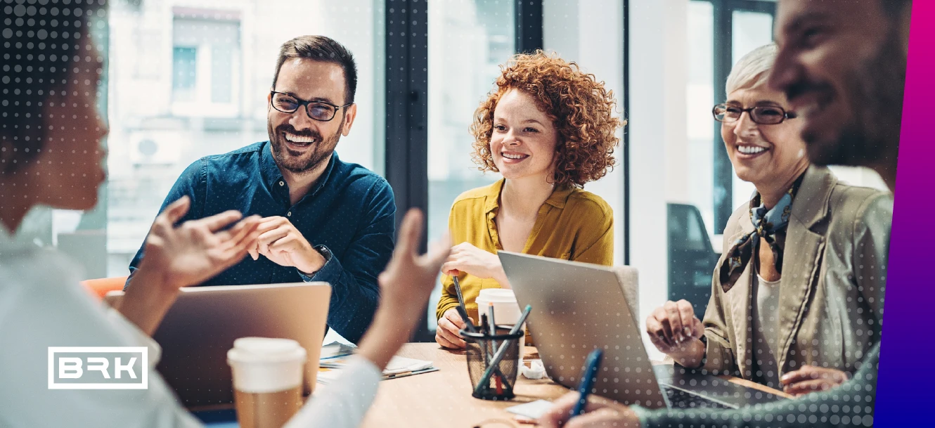 People smiling and talking around a table with laptops and notebooks in a meeting room.