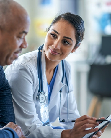Doctor with stethoscope talks with a patient, making notes on a clipboard, in a clinical setting.
