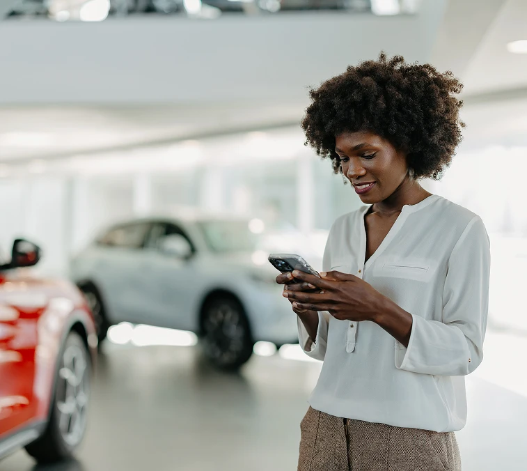 Woman with curly hair smiling while using her phone in a modern car showroom with cars in the background.