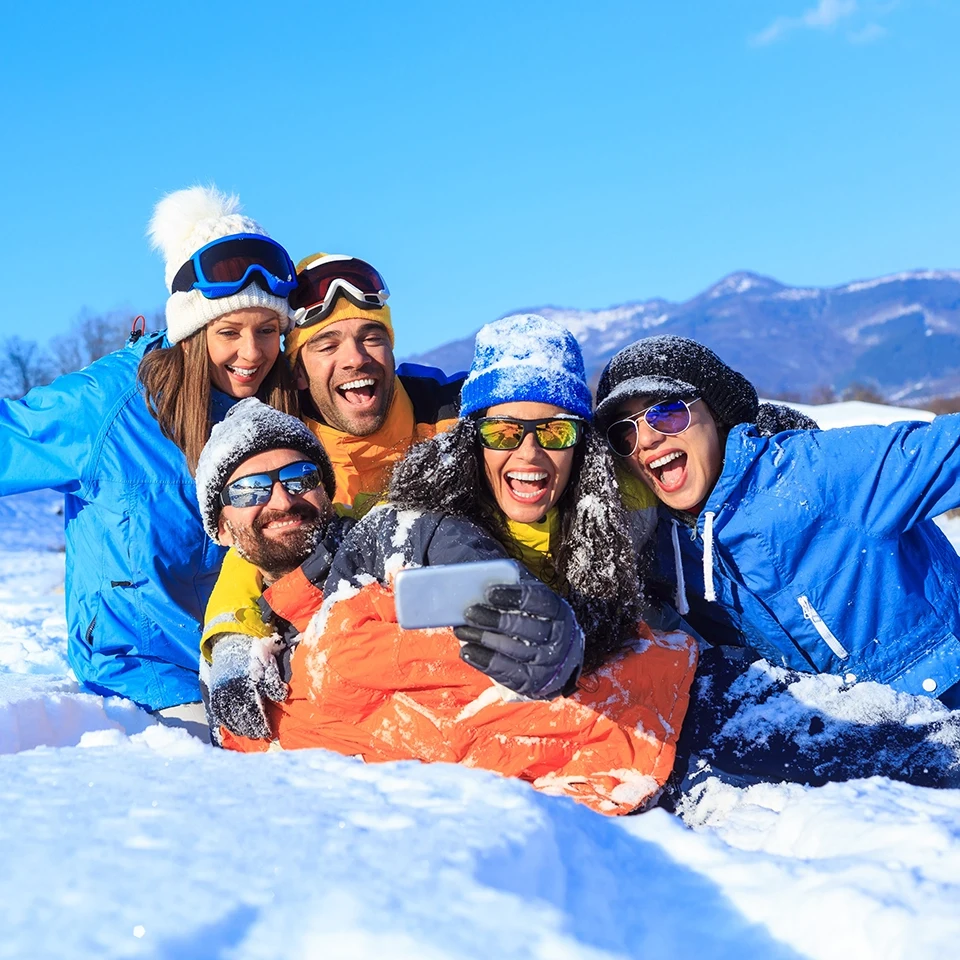 Five friends in colorful winter clothing smile and take a selfie while lying together on snowy ground outdoors.