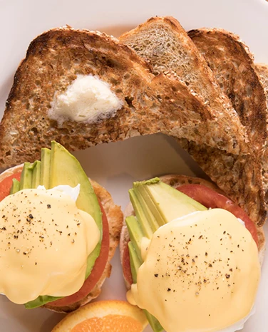 Toasted multigrain bread with butter, eggs benedict with avocado and tomato, and an orange slice on a plate.