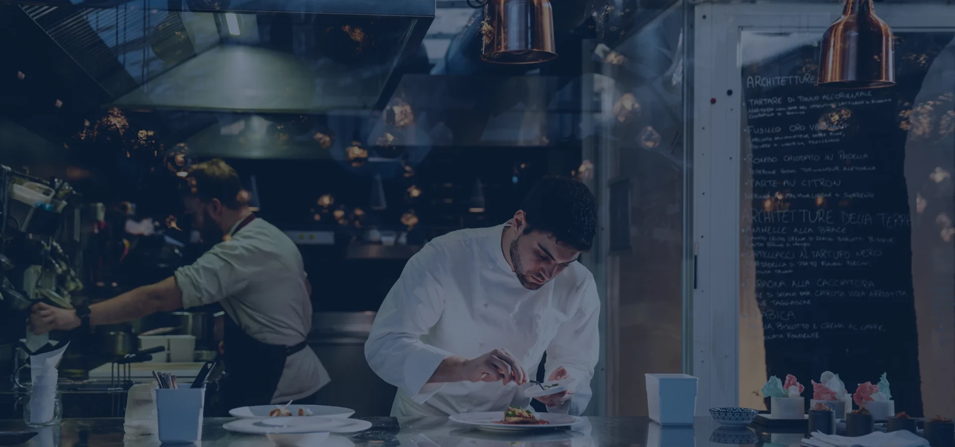 A chef carefully plating food in a restaurant kitchen, with another chef cooking in the background.