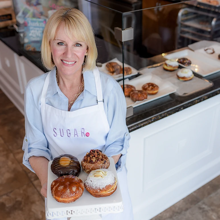 Woman in a bakery holding a tray of four assorted donuts, smiling at the camera.