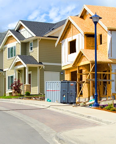 Two suburban houses, one completed and one under construction, on a residential street with a blue sky.
