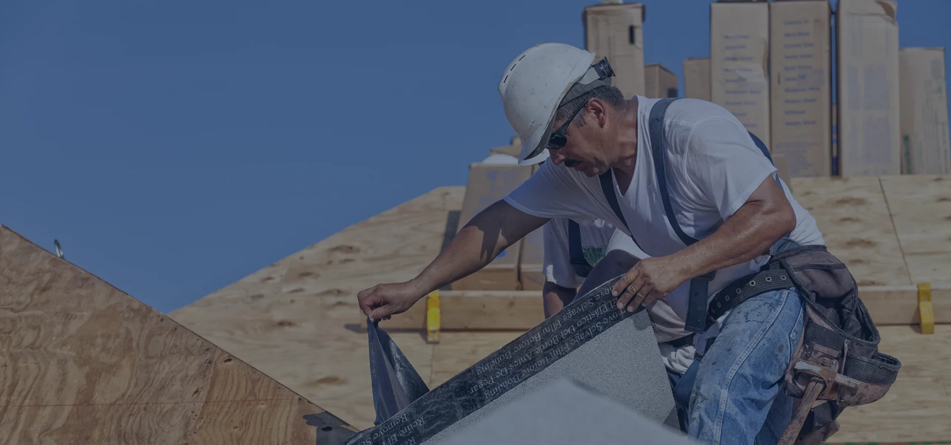 Construction worker in a hard hat installing shingles on a roof under a clear blue sky.