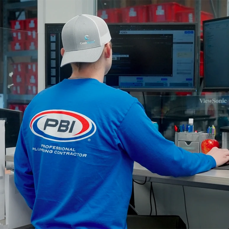 Person in a blue "PBI" shirt works at a computer desk with monitors and office supplies visible.