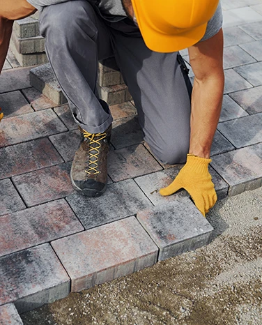 Worker in yellow gloves and boots laying concrete pavers on a pathway, kneeling on one knee.