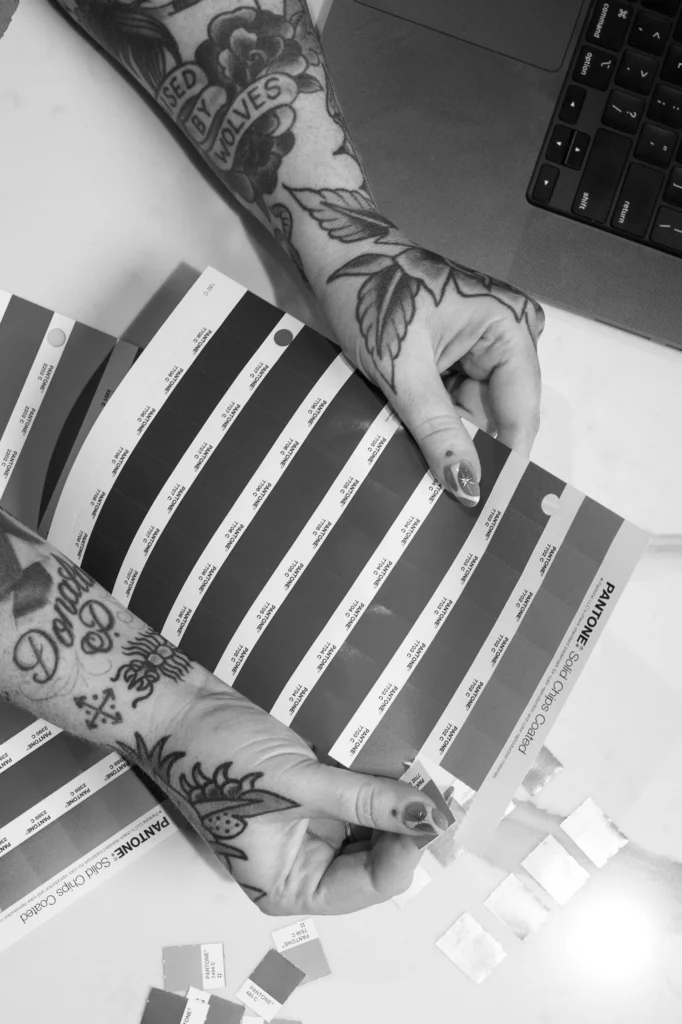Tattooed hands compare Pantone color swatches from a booklet next to a laptop on a white surface.