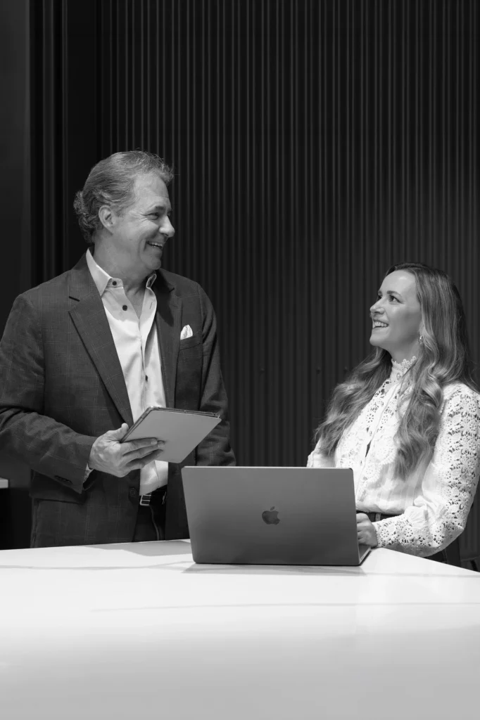 A man holding a tablet and a woman sitting at a table with a laptop smile at each other in an office setting.