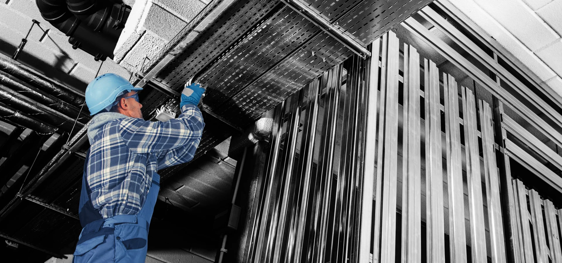Worker in blue helmet and overalls inspects or repairs cables on a ceiling in an industrial building.