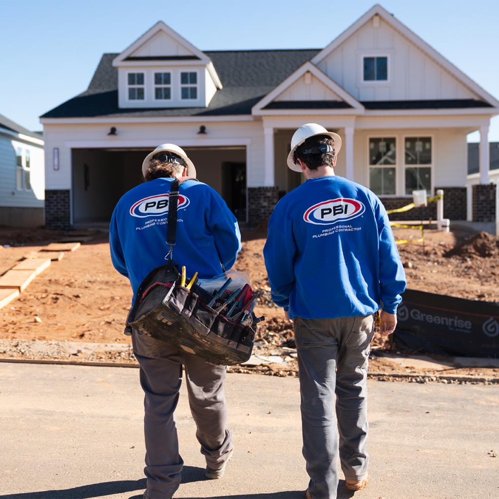 Two construction workers in blue shirts and helmets walk toward a new house under construction.