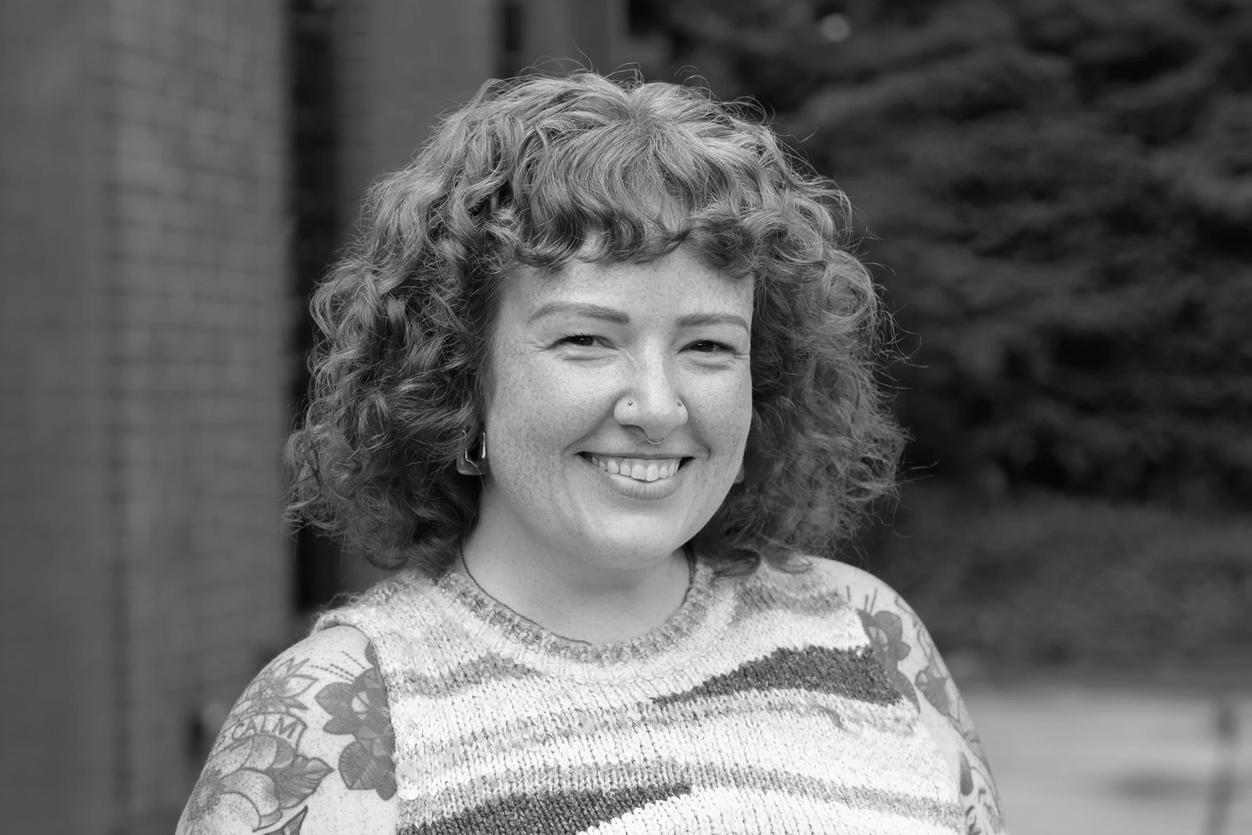 A person with curly hair and a nose piercing smiles at the camera, wearing a patterned sweater. The background, reminiscent of a Creative Agency’s vibrant outdoor space, shows brick and greenery.