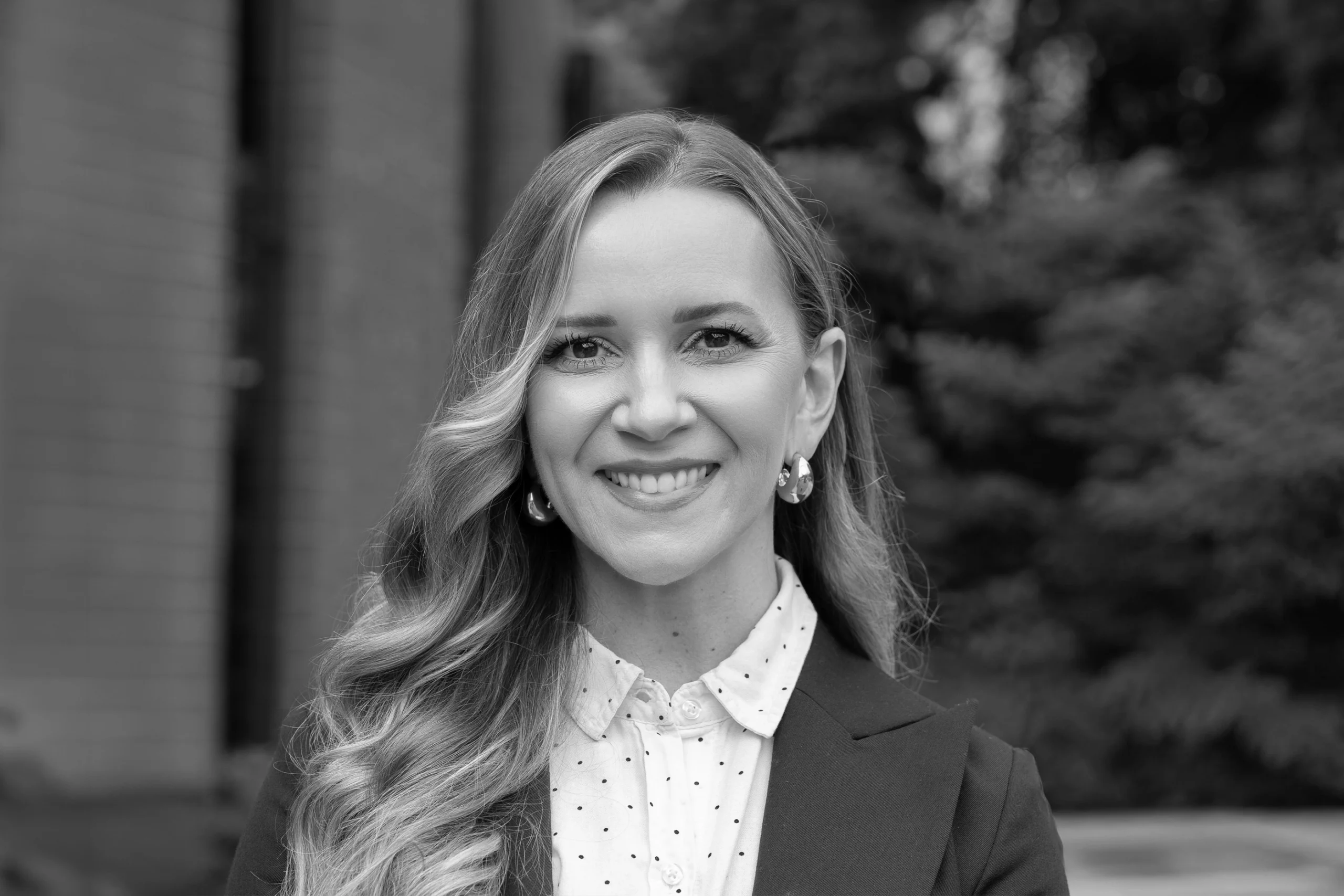 A woman with long wavy hair, wearing a blazer and polka dot shirt, smiles at the camera outdoors in front of a blurred building and greenery, embodying the confident spirit of a Creative Agency professional.