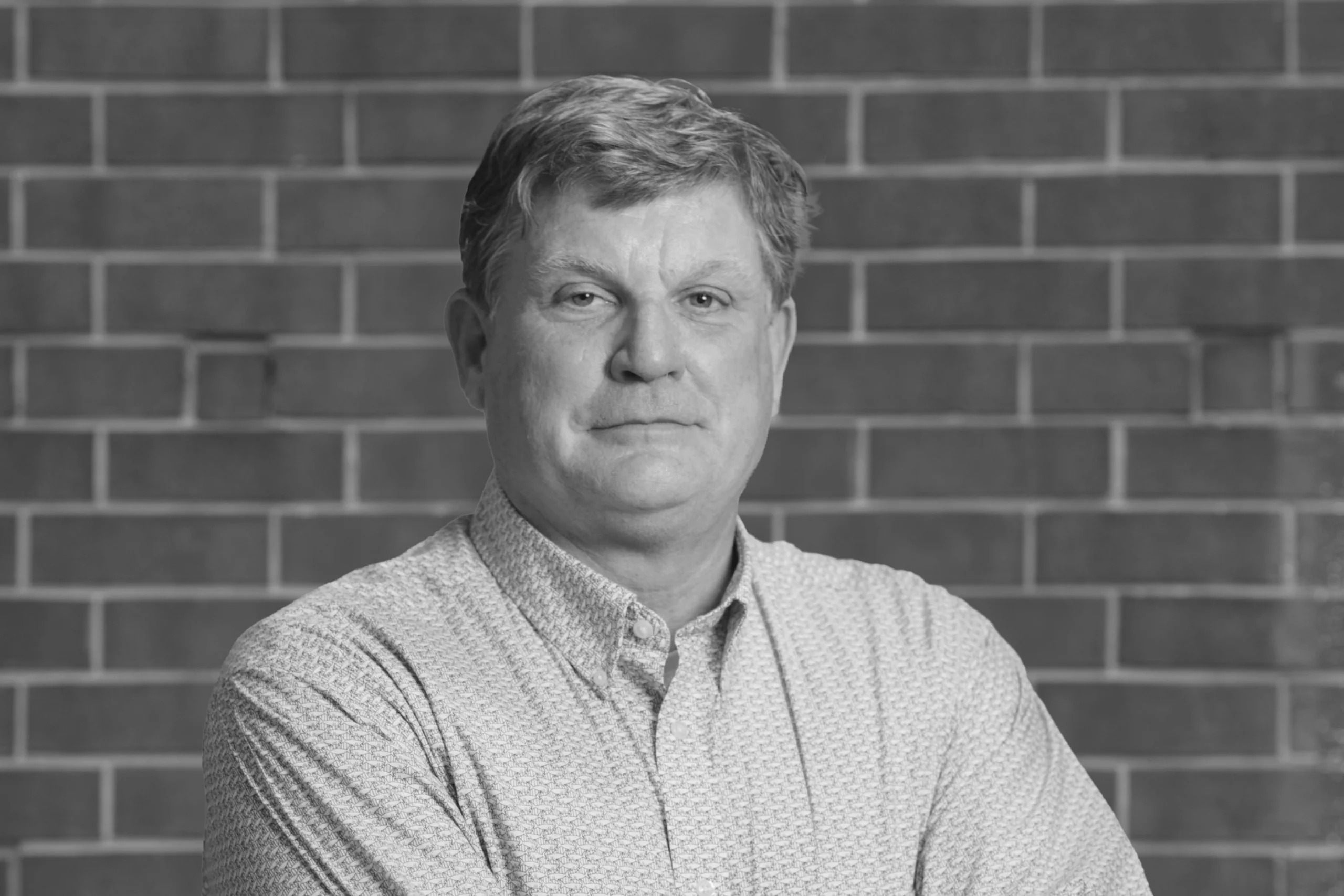 A middle-aged man with short hair, wearing a patterned shirt, stands in front of a brick wall with arms loosely crossed, looking at the camera. The image, captured in black and white, exudes the confident and thoughtful presence often seen in creative agency professionals.
