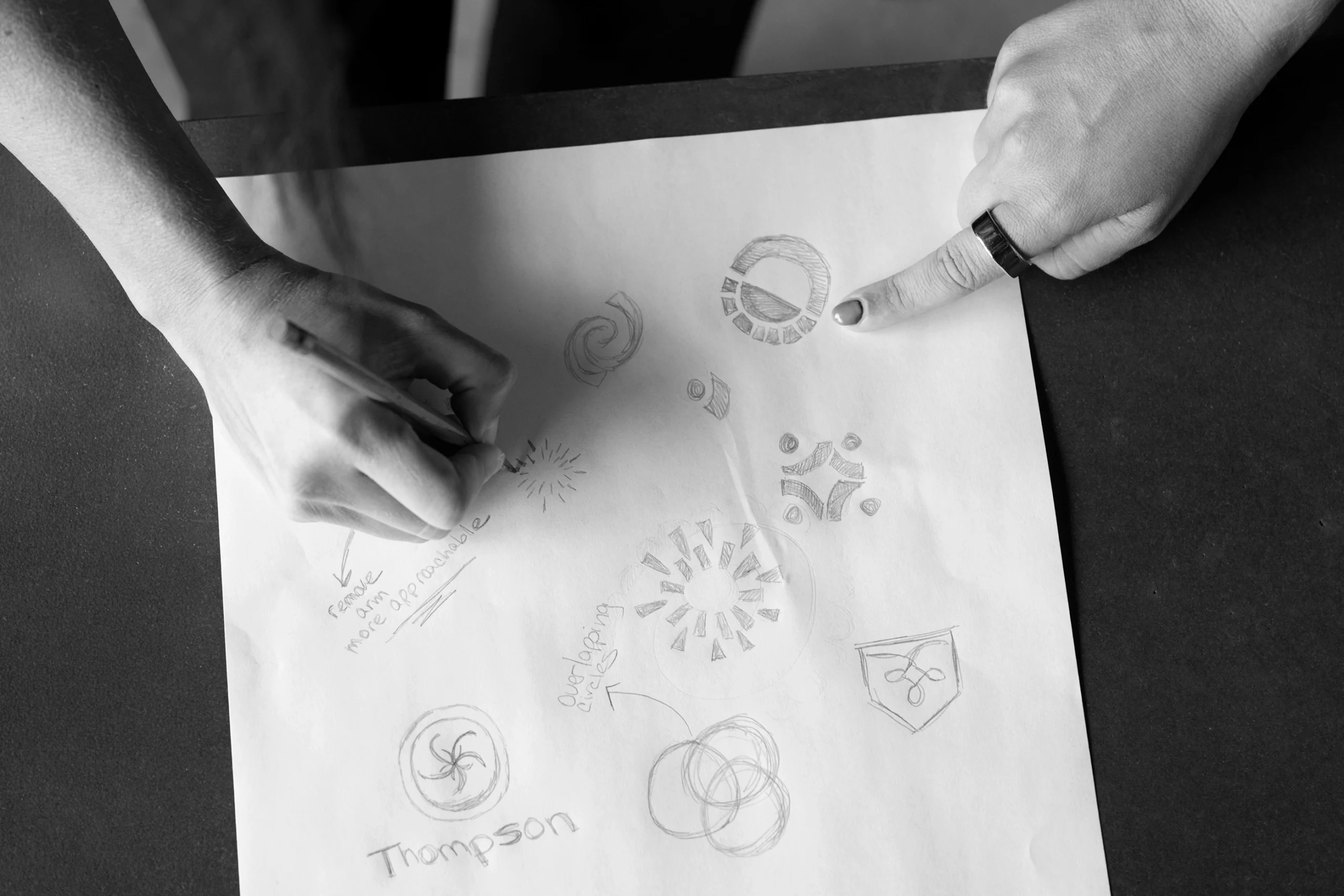 Two hands hover over a sheet of paper with multiple hand-drawn geometric sketches, circular patterns, and notes in pencil, capturing the imaginative process of a creative agency at work.