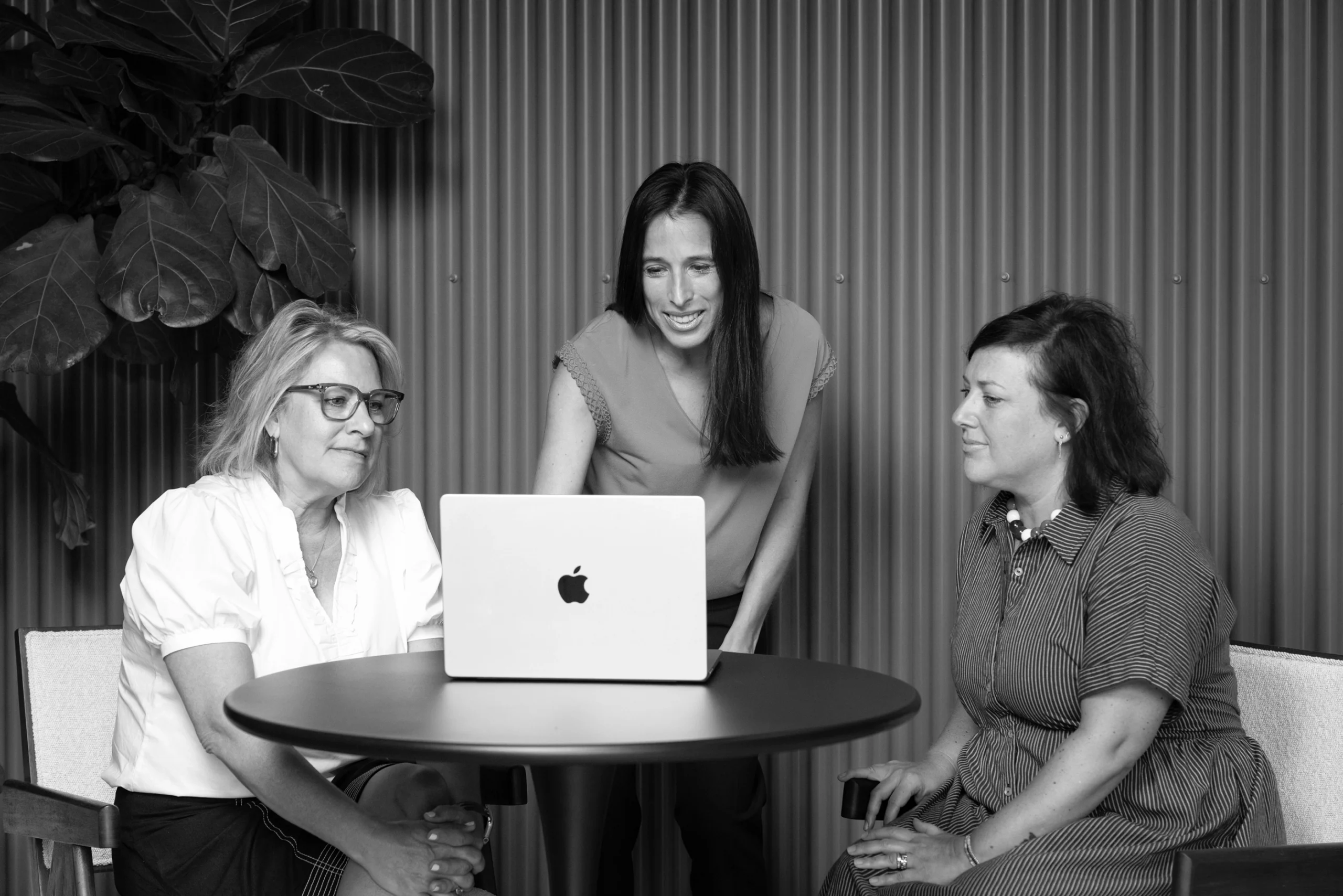 Three women sit and stand around a round table, looking at an open laptop together in a modern creative agency office setting.