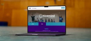 A laptop displays a website with the logo “hivemind” and navigation tabs including “purpose,” “habits,” and “the story,” set against a blurred indoor background.