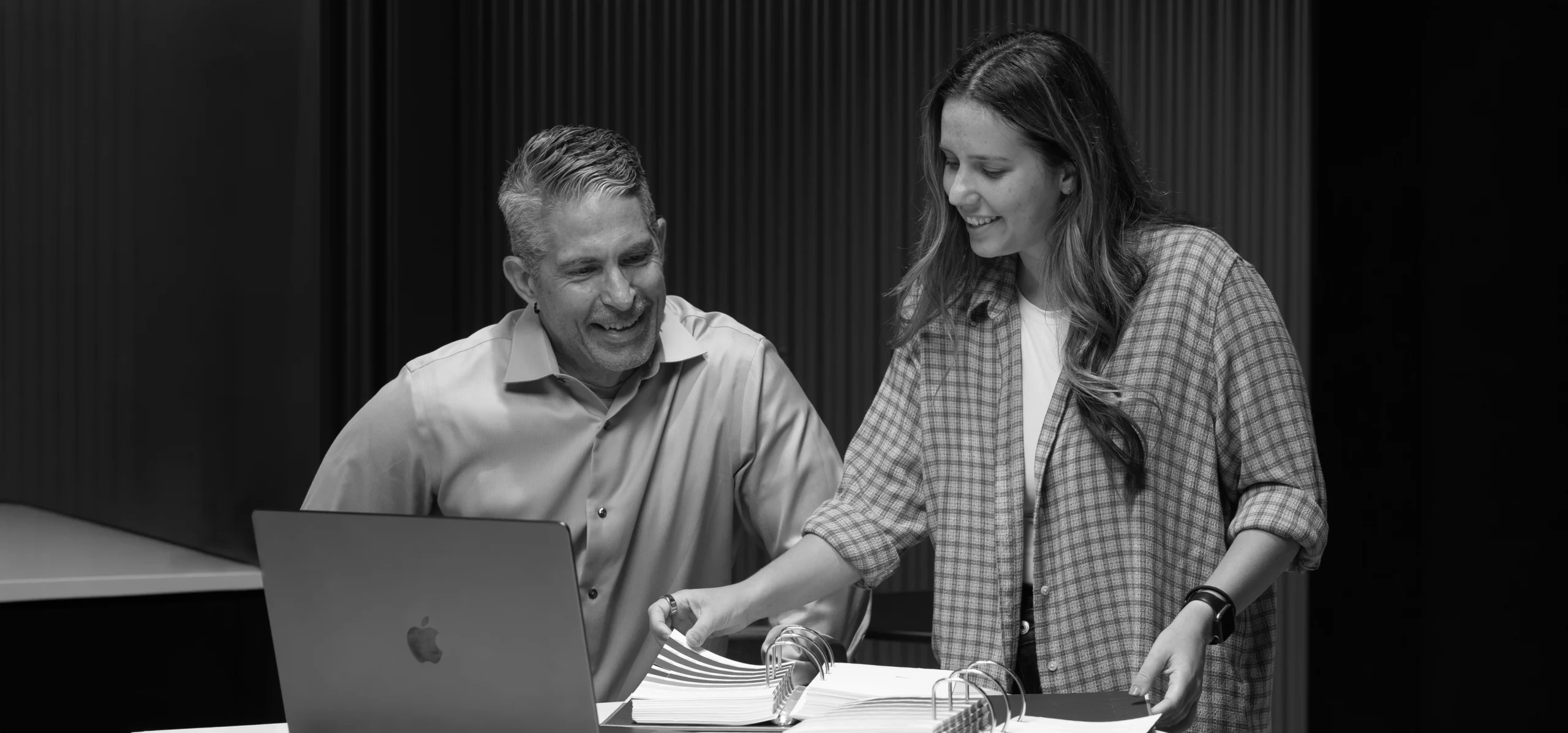 Two people standing at a table with a laptop and open books, smiling and discussing documents in a modern office setting.