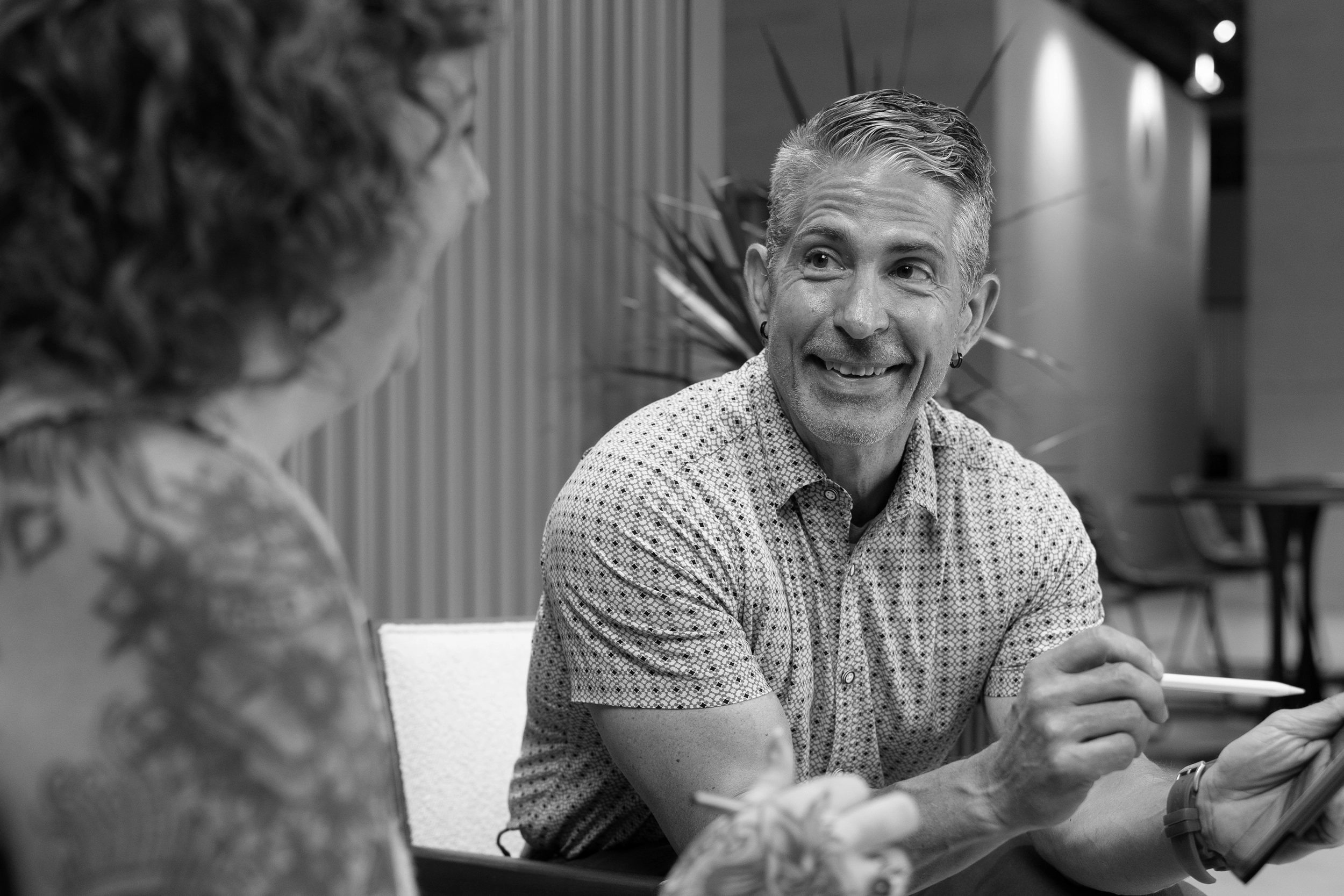 A man smiles and gestures with a pen while speaking to a woman in a modern indoor setting, capturing the dynamic and collaborative atmosphere often found in a Creative Agency. The image is in black and white.