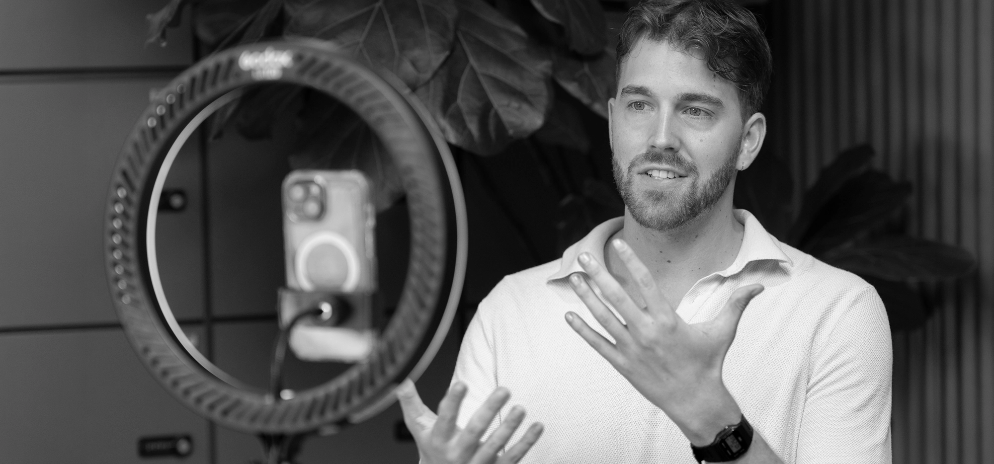 A man gestures while speaking in front of a smartphone mounted on a ring light, appearing to record or livestream indoors.
