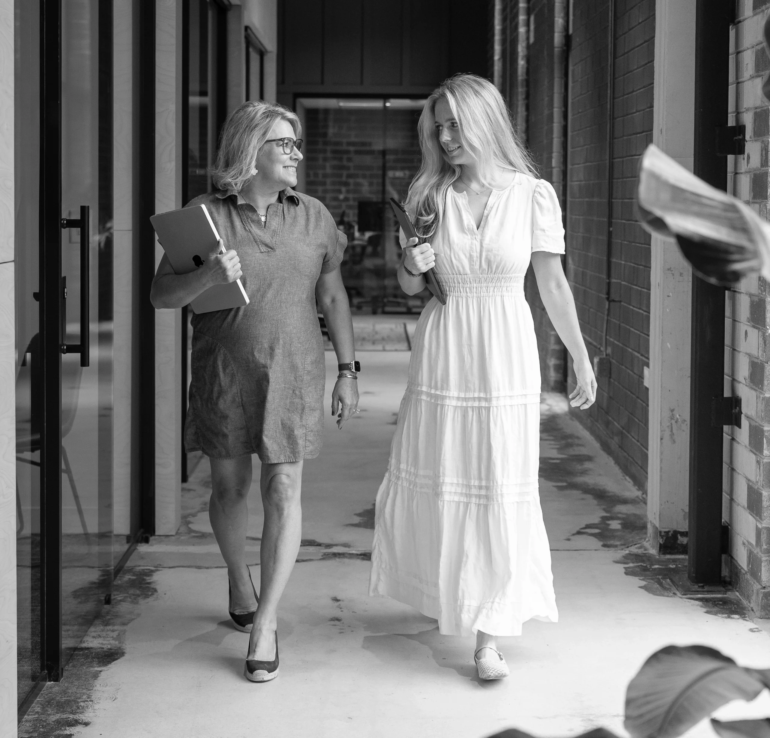 Two people walk together in a hallway; one holds a laptop and wears glasses, the other wears a long white dress. The scene is in black and white.