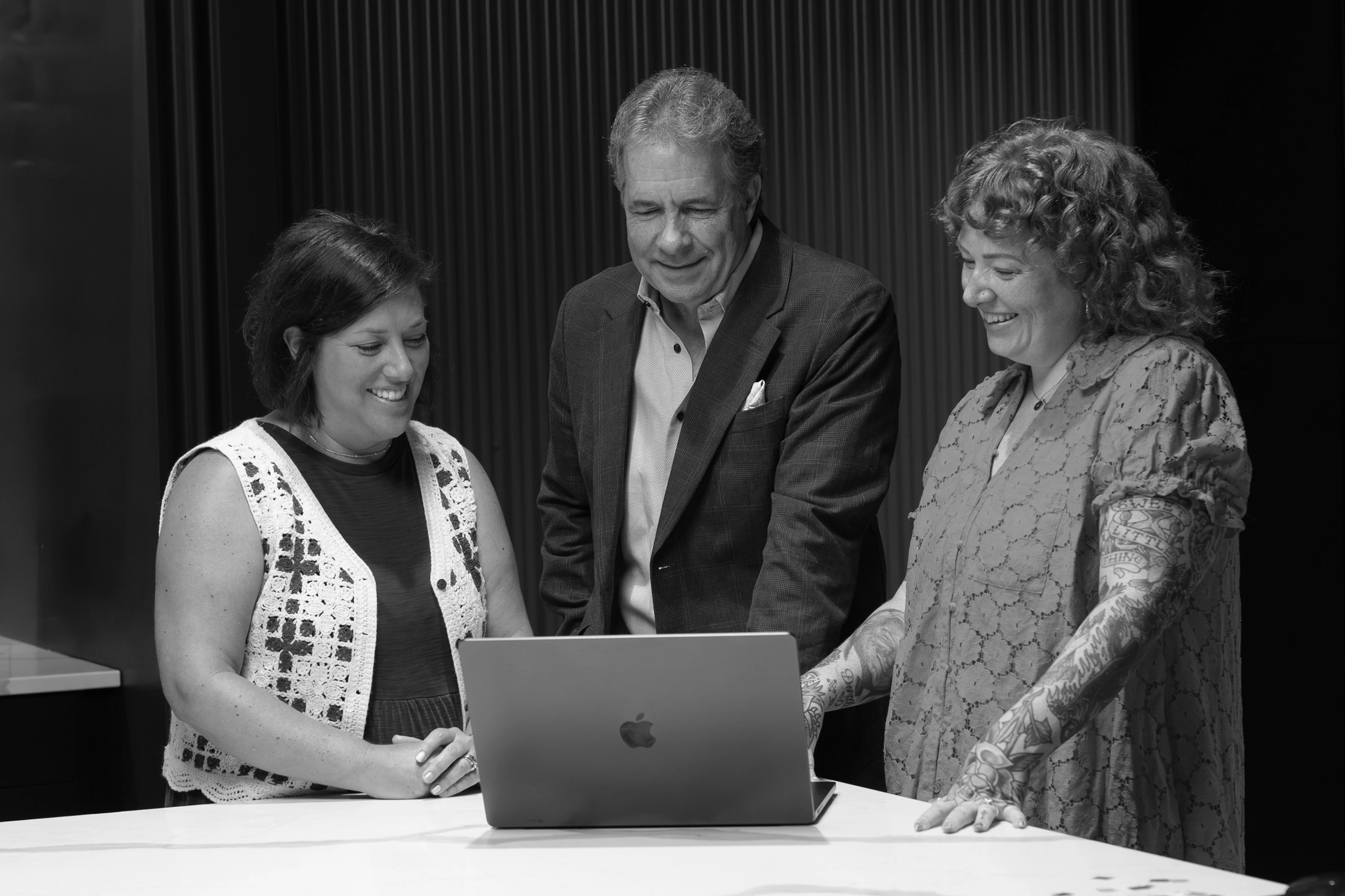 Three people stand around a table, looking at a laptop and smiling, embodying the collaborative spirit of a creative agency. The photo is in black and white, with a plain background.