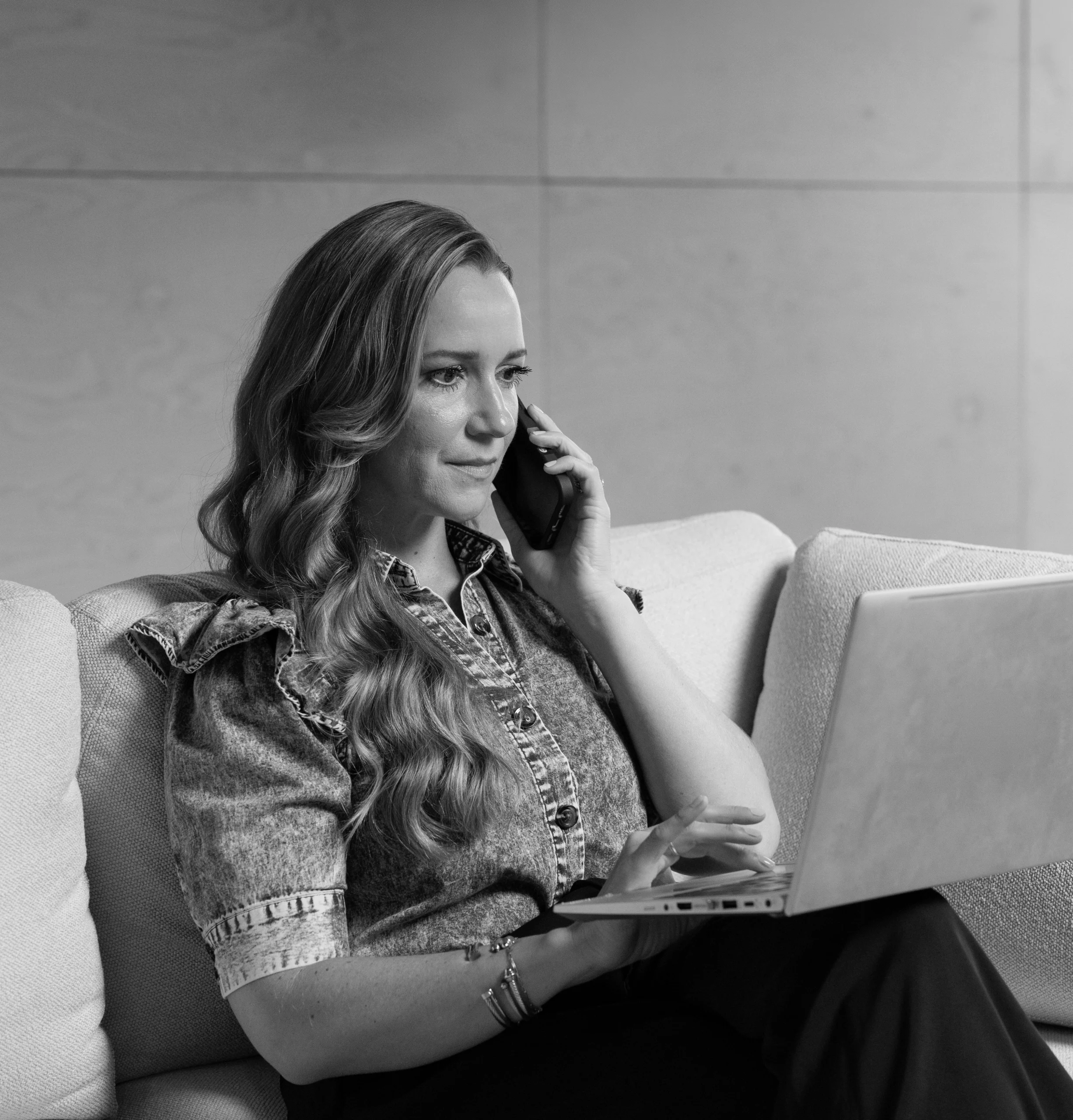 A woman sits on a couch, talking on a smartphone while using a laptop. She appears focused and is dressed in a ruffled shirt.