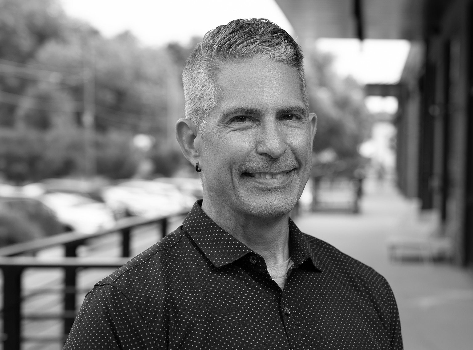 A man with short, gray hair and an earring smiles outdoors, standing on a sidewalk next to a metal railing. The background is blurred with trees and parked cars visible, capturing the casual confidence often seen in professionals from a creative agency.