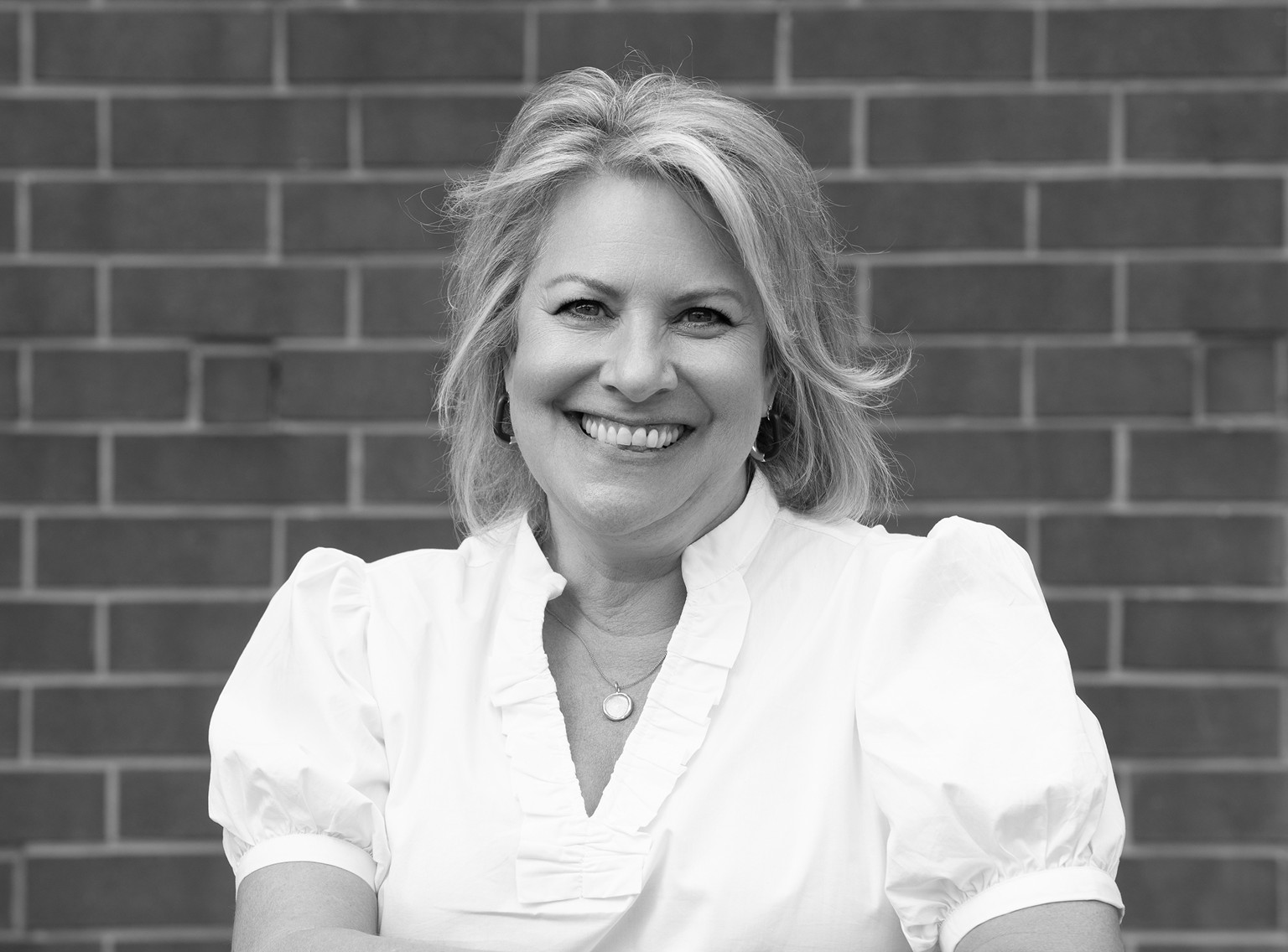 Smiling woman with light hair wearing a white blouse, standing in front of a brick wall, exuding the confident and approachable vibe of a Creative Agency professional.
