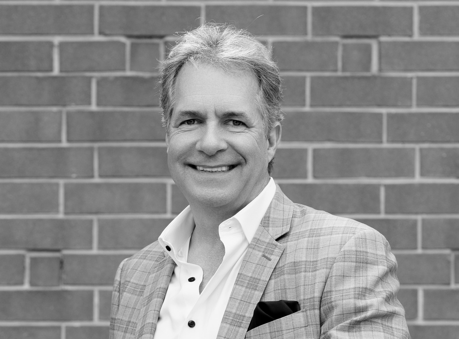 Smiling man in a light plaid blazer and open-collared white shirt stands in front of a brick wall, exuding the approachable professionalism of a creative agency.